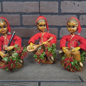 May include: Three vintage Christmas elf figurines. Each elf wears a red outfit and hat, with gold accents. One elf holds a flute, another a lute, and the third plays drums. Each is surrounded by a wreath of red flowers and green leaves.