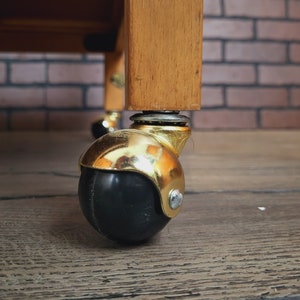May include: A close-up of a gold caster wheel with a black rubber tyre. The wheel is attached to a wooden table leg.