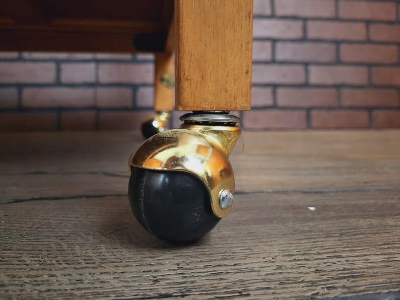 May include: A close-up of a gold caster wheel with a black rubber tyre. The wheel is attached to a wooden table leg.