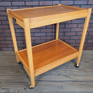 May include: A wooden two-tiered serving trolley with wheels. The trolley has a natural wood finish and is made of solid wood. The trolley has a drawer on the top shelf.