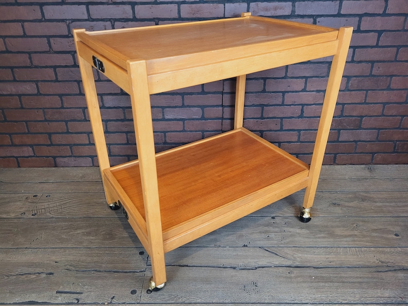 May include: A wooden two-tiered serving trolley with wheels. The trolley has a natural wood finish and is made of solid wood. The trolley has a drawer on the top shelf.