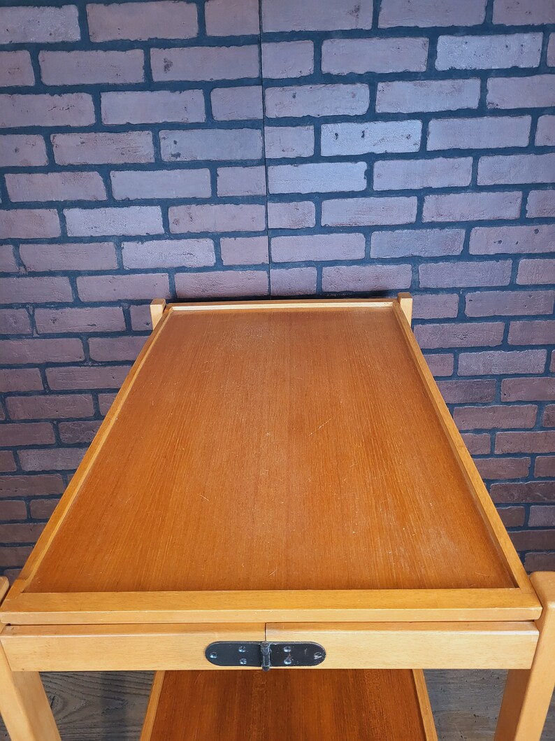 May include: A wooden serving trolley with two shelves and a black metal hinge. The trolley is made of light wood and has a natural finish.