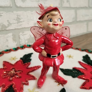 May include: A red and white ceramic Christmas elf figurine with pink wings. The elf is wearing a red hat, a red suit with a black belt, and has a smiling face.