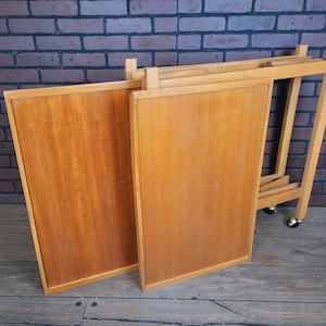 May include: A wooden folding serving trolley with four wheels. The trolley is made of light wood and has a natural finish. The trolley is folded up and ready to be stored.