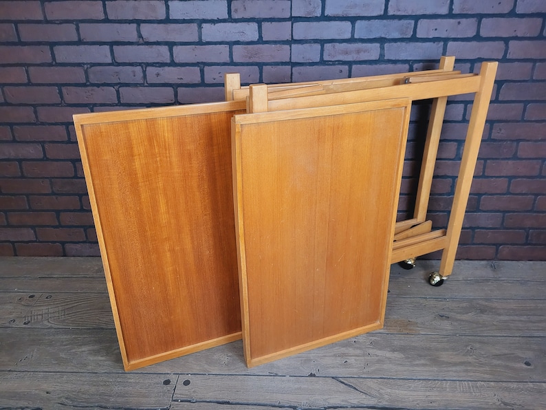 May include: A wooden folding serving trolley with four wheels. The trolley is made of light wood and has a natural finish. The trolley is folded up and ready to be stored.