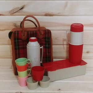 May include: A red and black plaid picnic basket with a brown leather handle, a white and red thermos, and six colorful plastic cups.