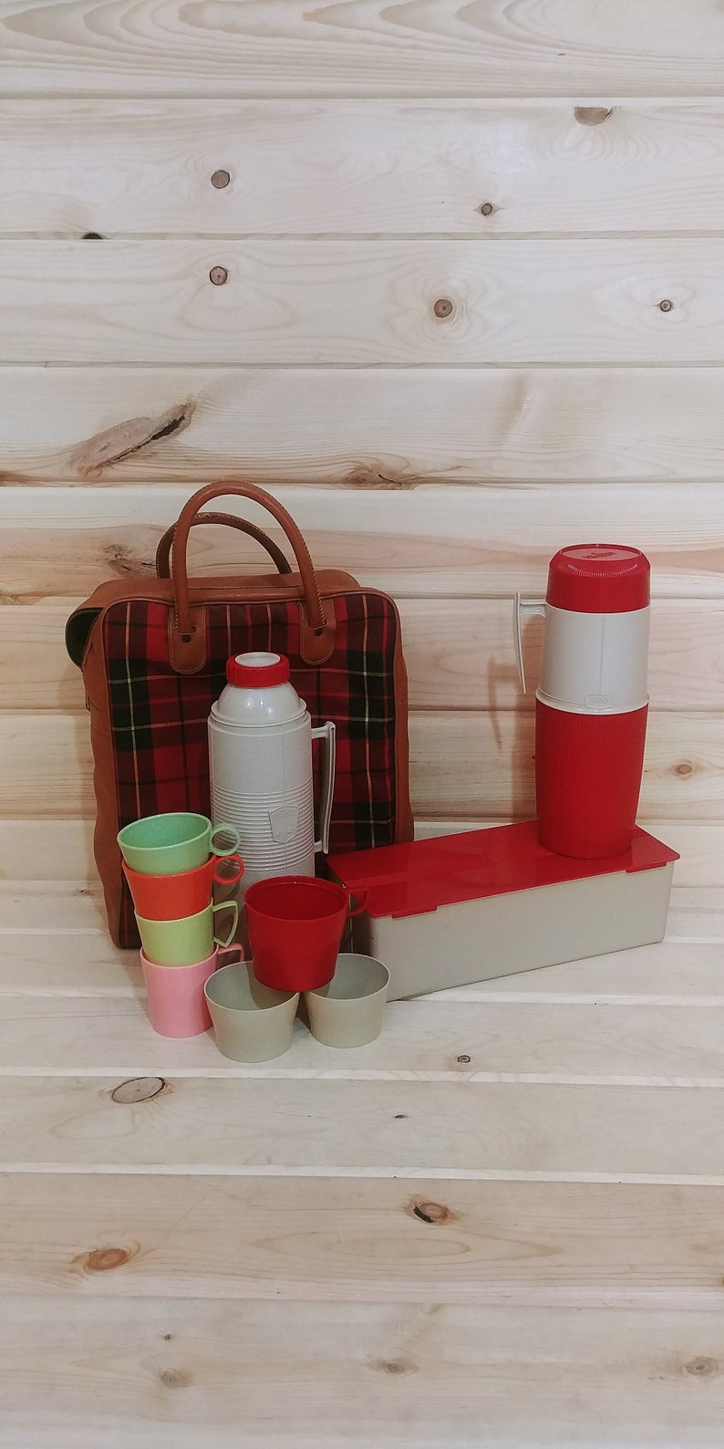 May include: A red and black plaid picnic basket with a brown leather handle, a white and red thermos, and six colorful plastic cups.