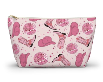 Peut inclure: Pochette cosmétique rose avec un motif répété de chapeaux de cow-boy roses, de bottes et de boules à facettes sur un fond rose clair.