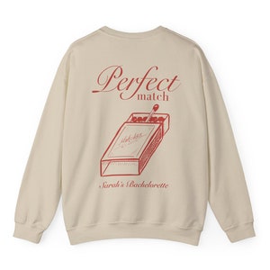 May include: A beige sweatshirt with a red graphic of a matchbox and the words "Perfect match" and "Sarah's Bachelorette". The matchbox design includes the words "Matches C-matic".
