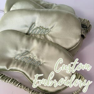 May include: Several sage green sleep masks with custom embroidery. The masks have an elastic band and are made of a silky material. The words "lane" and "dano" are embroidered on the masks. The text "Custom Embroidery" is also visible.