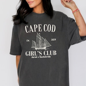 Puede incluir: Una camiseta gris oscuro con un diseño gráfico blanco. El diseño presenta un velero, el texto "CAPE COD", "Est. 2024", "GIRL'S CLUB" y "Sarah's Bachelorette".