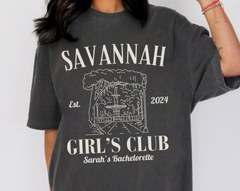 Può includere: Una maglietta grigio scuro con un design grafico bianco. Il grafico presenta un disegno di una fontana in un parco con alberi e lampioni. Il testo "Savannah Est. 2024 Girl's Club Sarah's Bachelorette" è stampato sulla maglietta.