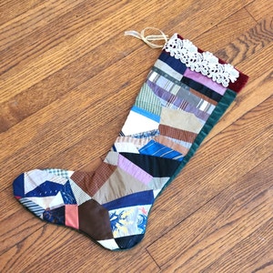 May include: A handmade Christmas stocking with a patchwork design. The stocking features various fabric patterns in shades of blue, brown, and purple. The top is trimmed with white lace and a burgundy velvet border. A cream-colored ribbon is attached for hanging.