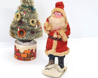 May include: Vintage Christmas decoration set featuring a Santa Claus figurine with a red suit, white beard, and a white fur trim. The Santa is standing on a white base with a brown brick chimney. A small, green, flocked Christmas tree with red and blue ornaments is also included. The base of the tree has a brick chimney design and says "Japan" and "On" on the bottom.