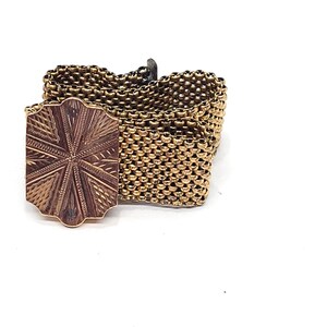May include: A gold-toned chain bracelet with a decorative, patterned clasp. The clasp is a square shape with a starburst design.