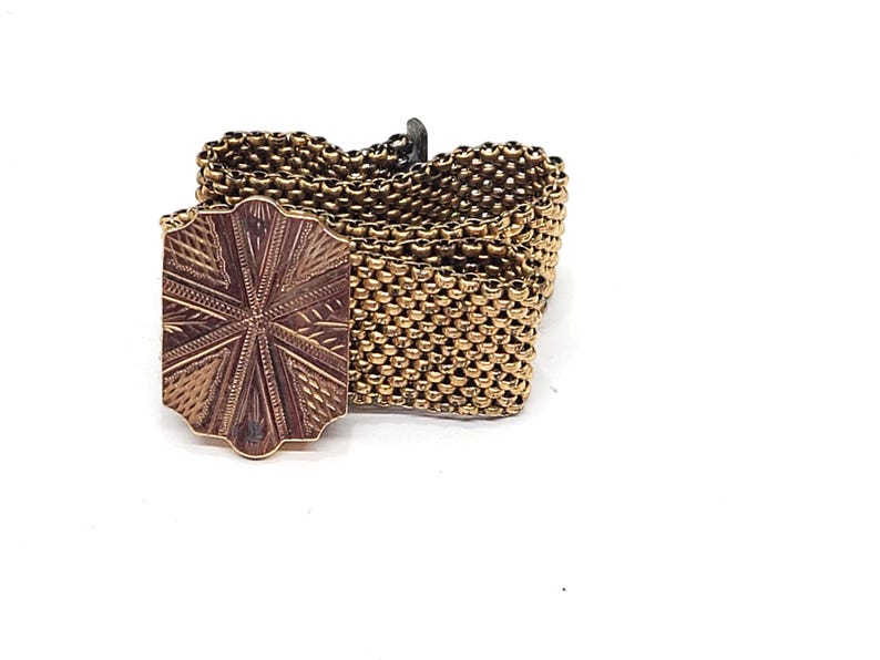 May include: A gold-toned chain bracelet with a decorative, patterned clasp. The clasp is a square shape with a starburst design.