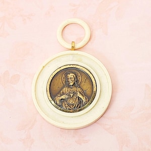 May include: A white plastic pendant with a gold-toned image of Jesus Christ with a heart on his chest. The pendant has a ring at the top for hanging.