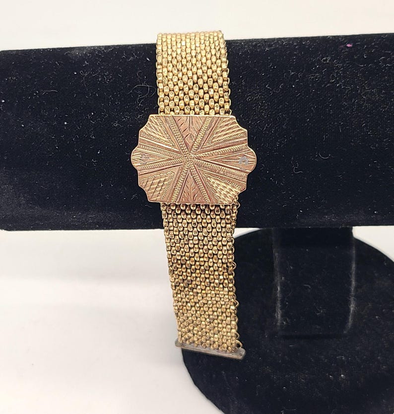 May include: A gold-toned chain bracelet with a decorative, engraved clasp. The bracelet is made of a woven chain pattern.