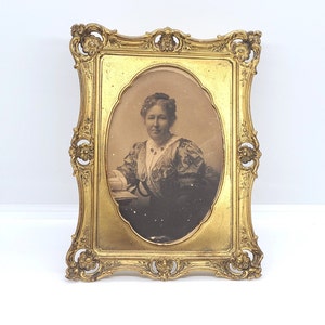May include: A vintage gold-toned ornate frame with a black and white photo of a woman wearing a patterned dress and a pearl necklace.