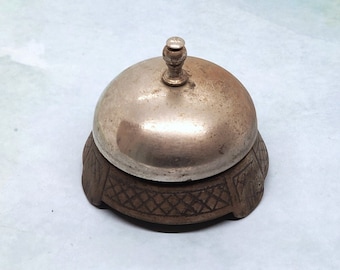 May include: A silver bell with a decorative base. The bell has a smooth, rounded top and a small knob on top. The base is made of a dark metal with a textured design.