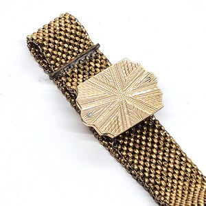May include: A gold mesh bracelet with a decorative clasp. The clasp is a rectangular shape with a starburst design.