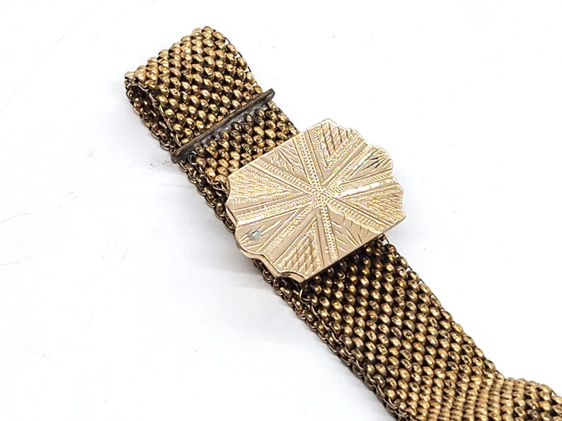 May include: A gold mesh bracelet with a decorative clasp. The clasp is a rectangular shape with a starburst design.