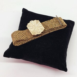 May include: A gold-toned metal bracelet with a decorative, patterned clasp. The bracelet is on a black velvet surface.