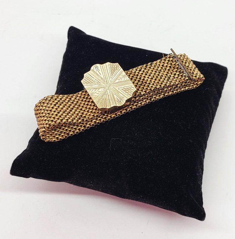 May include: A gold-toned metal bracelet with a decorative, patterned clasp. The bracelet is on a black velvet surface.