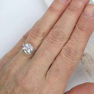 May include: A silver ring with a large, clear, round gemstone set in a claw setting. The ring is on a person's finger.