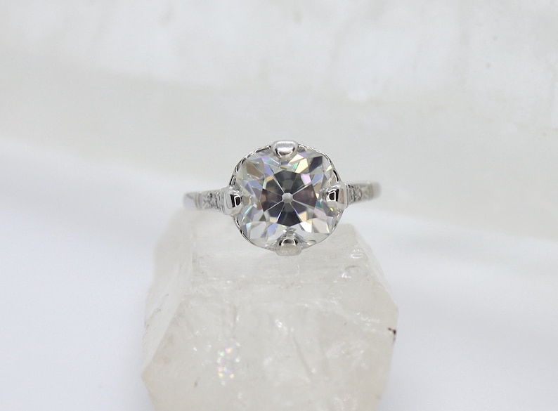 May include: A silver ring with a large, clear, round gemstone set in a delicate, ornate setting. The ring is on a white background.