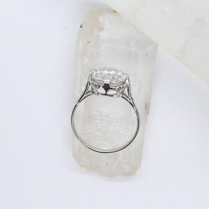 May include: A silver ring with a large, clear, round gemstone set in a delicate, ornate setting. The ring is displayed on a white crystal.