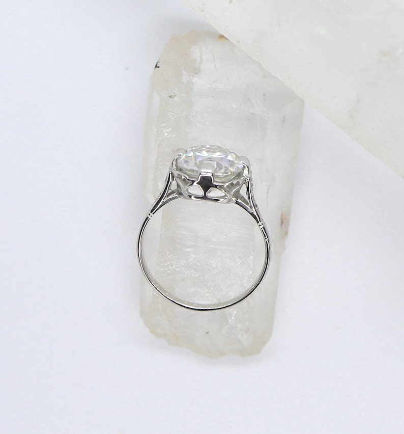 May include: A silver ring with a large, clear, round gemstone set in a delicate, ornate setting. The ring is displayed on a white crystal.