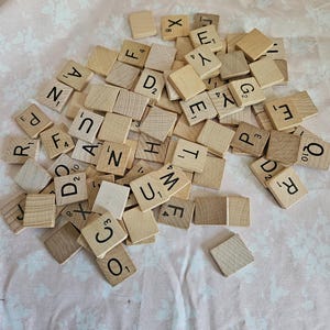 May include: A pile of wooden letter tiles, commonly used in the game Scrabble, are scattered on a light pink fabric with a floral pattern. The tiles are all in various positions, with some letters facing up and others facing down.