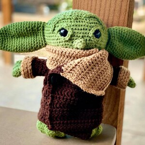 May include: A crocheted green and brown plush toy with large ears and a brown scarf. The toy is wearing a brown coat and has large black eyes.