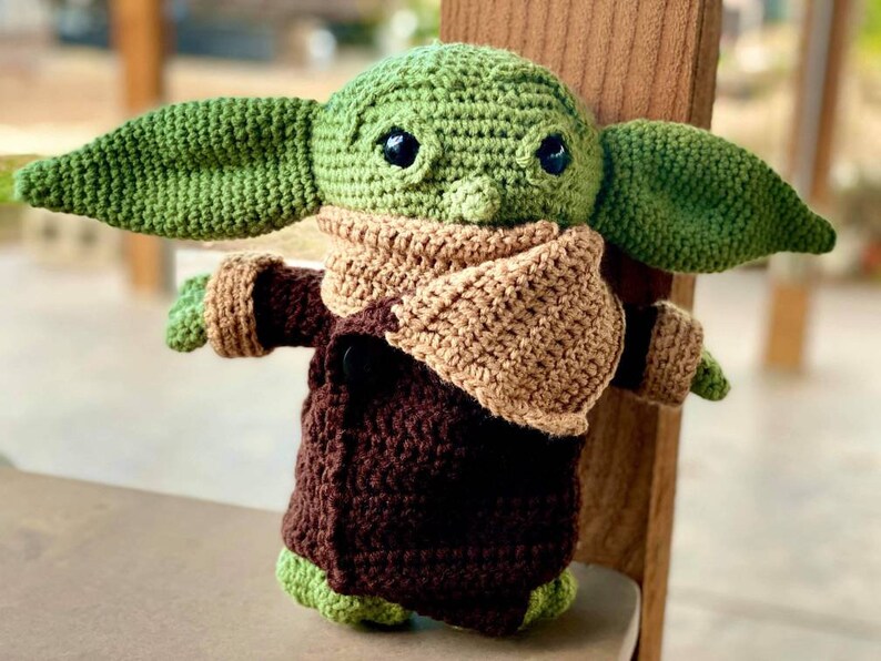 May include: A crocheted green and brown plush toy with large ears and a brown scarf. The toy is wearing a brown coat and has large black eyes.