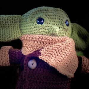 May include: A crocheted plush toy resembling a baby alien with large ears, wearing a brown coat and a pink scarf. The toy has large, dark eyes and a button on its coat.