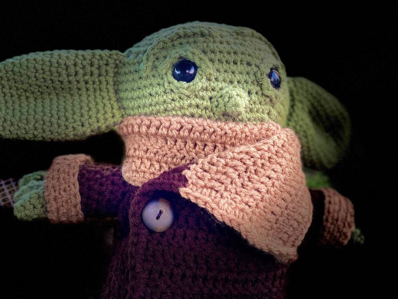 May include: A crocheted plush toy resembling a baby alien with large ears, wearing a brown coat and a pink scarf. The toy has large, dark eyes and a button on its coat.