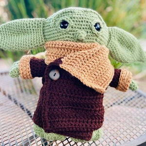 May include: A crocheted green and brown plush toy resembling a character from the Star Wars franchise. The toy is wearing a brown coat with a tan collar and has large eyes.