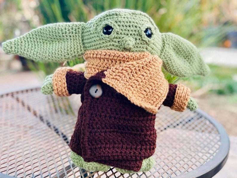May include: A crocheted green and brown plush toy resembling a character from the Star Wars franchise. The toy is wearing a brown coat with a tan collar and has large eyes.
