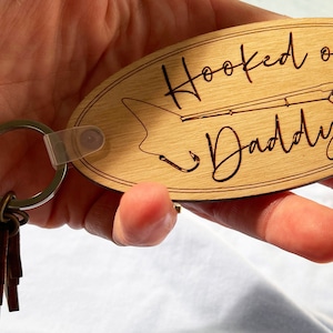 May include: Wooden oval keychain with the text "Hooked on Daddy" and a fishing rod design. The keychain is light brown with a metal key ring and several small brown leather tags.