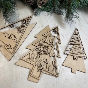 May include: Three light-colored wooden Christmas tree ornaments with laser-cut designs. One features a truck carrying a tree, another displays the text Let it snow with a snowflake, and the third has a simple geometric line pattern.