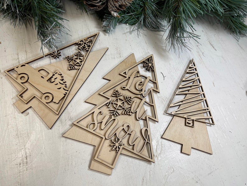May include: Three light-colored wooden Christmas tree ornaments with laser-cut designs. One features a truck carrying a tree, another displays the text Let it snow with a snowflake, and the third has a simple geometric line pattern.