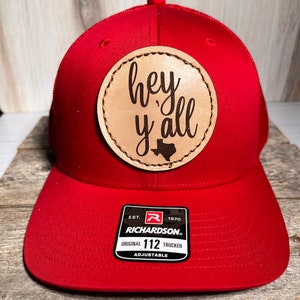 May include: A bright red trucker hat featuring a circular tan leather patch centered on the front. The patch is laser-engraved with the text "hey y'all" and a small silhouette of the state of Texas. A black and white sticker on the brim reads "RICHARDSON ORIGINAL 112 TRUCKER ADJUSTABLE".