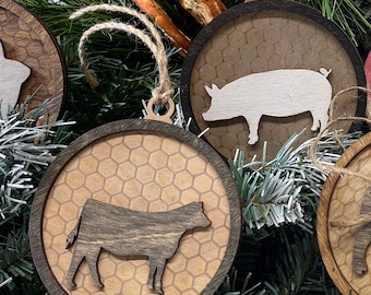 May include: Wooden Christmas ornaments with farm animal silhouettes. Each round ornament features a laser-cut animal, including a cow and a pig, set against a honeycomb pattern. The ornaments are trimmed with a dark wood border and have twine hangers.