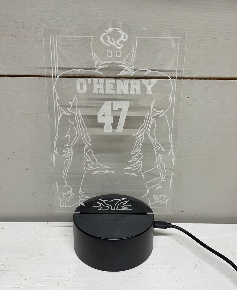 May include: A clear acrylic LED lamp with a football player design. The player's jersey shows the name "O'Henry" and the number "47" in white. The lamp is mounted on a black base with a power cord.