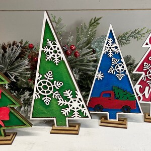 May include: Four decorative wooden Christmas tree cutouts standing on small bases. One green tree features red bows, another green tree has white snowflakes, a blue tree displays a red truck, and a red tree features white snowflakes and the text Let it snow.