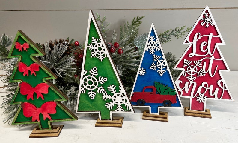 May include: Four decorative wooden Christmas tree cutouts standing on small bases. One green tree features red bows, another green tree has white snowflakes, a blue tree displays a red truck, and a red tree features white snowflakes and the text Let it snow.