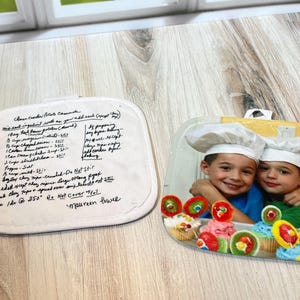 May include: Two square pot holders. One features a handwritten recipe for Cheese Cracker Potato Casserole signed by Maureen Power. The other displays a photograph of two children wearing white chef hats, smiling behind a tray of colorful cupcakes.