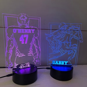 May include: Two illuminated acrylic night lights. One features a football player with the name "O'HENRY" and the number "47". The other depicts a jockey on a horse with the name "GABBY". Both lights sit on black bases.