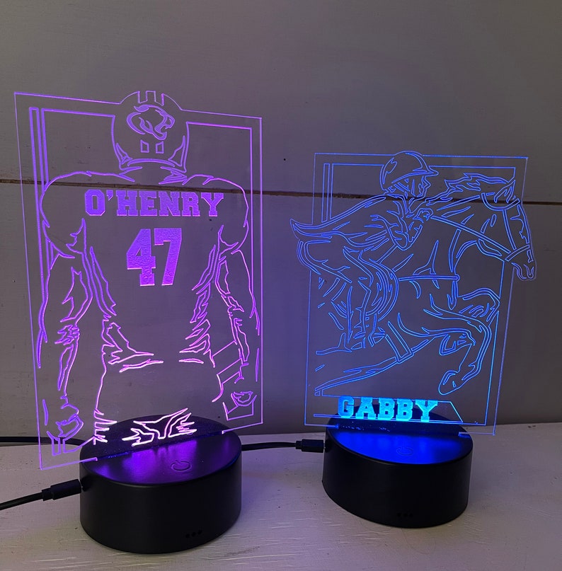 May include: Two illuminated acrylic night lights. One features a football player with the name "O'HENRY" and the number "47". The other depicts a jockey on a horse with the name "GABBY". Both lights sit on black bases.